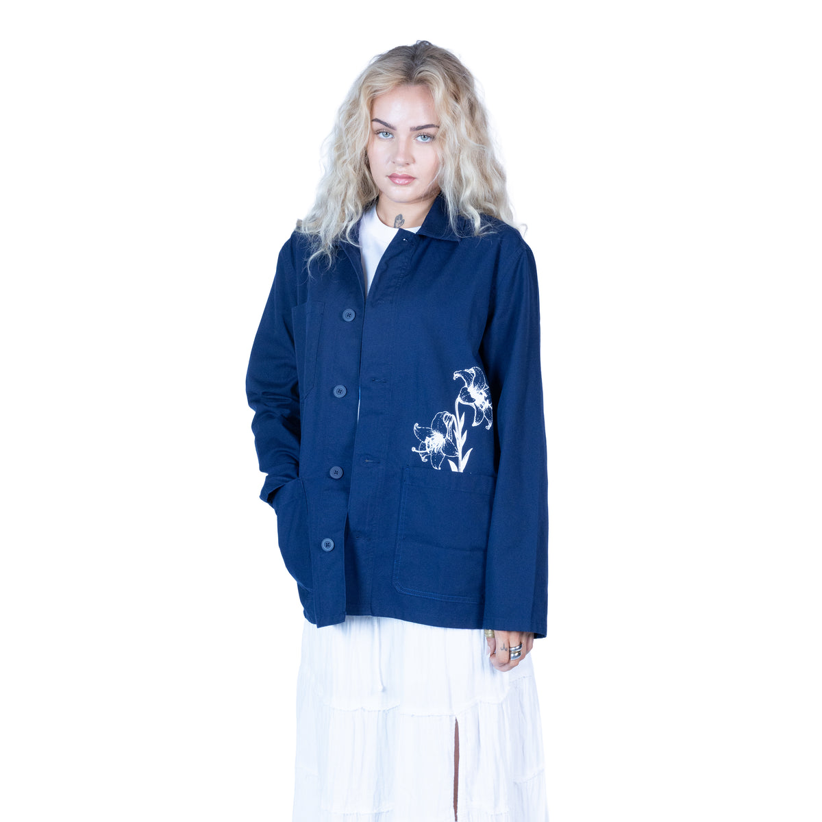 A model with blonde hair wearing a navy blue button-up jacket with white floral embroidery on the pocket, paired with a white top and white skirt, against a solid white background.