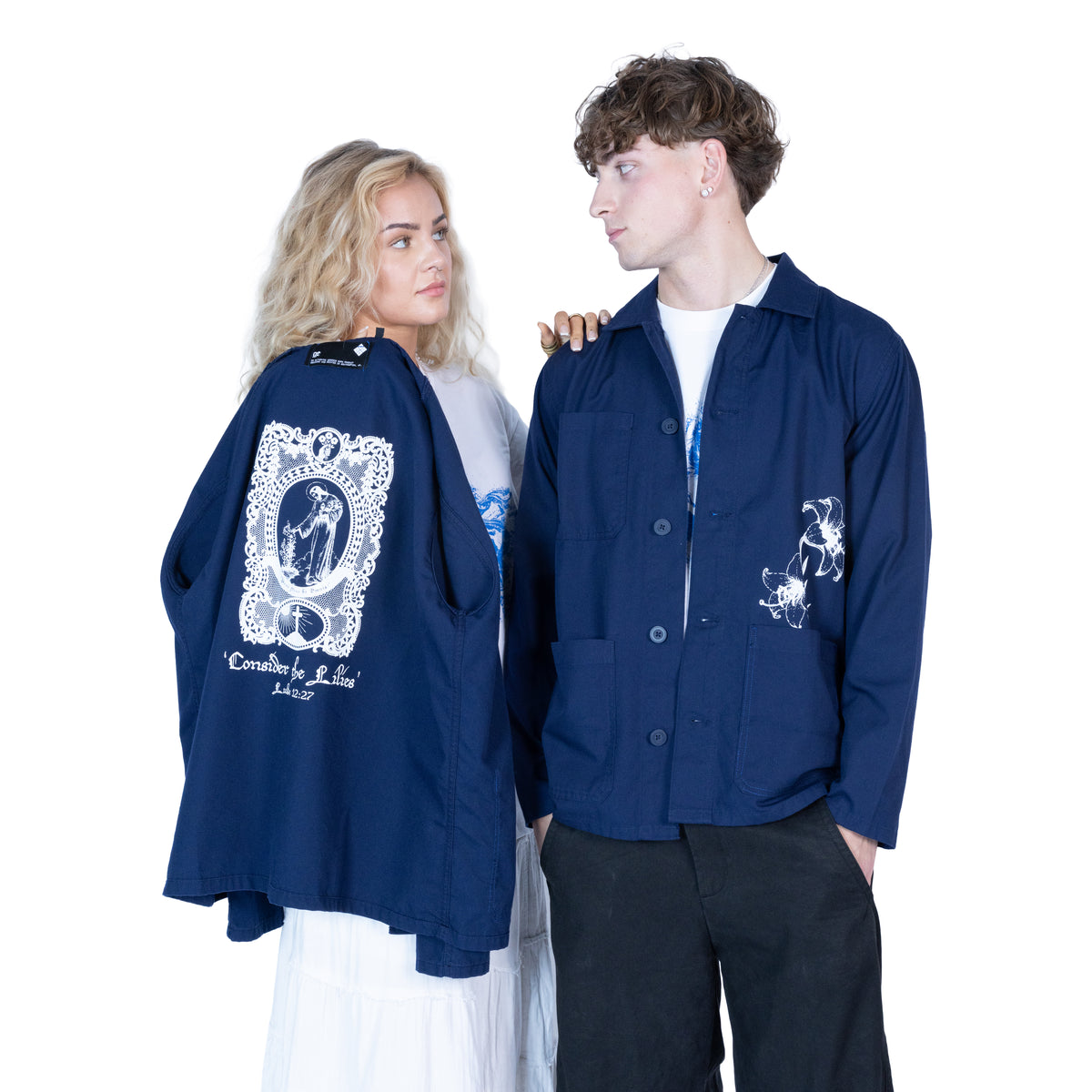 A man and a woman modeling navy blue work shirts with white graphic prints. The woman holds up the back of the shirt, which features a large white religious illustration and the text 'Consider the Lilies' from the bible verse Luke 12:27