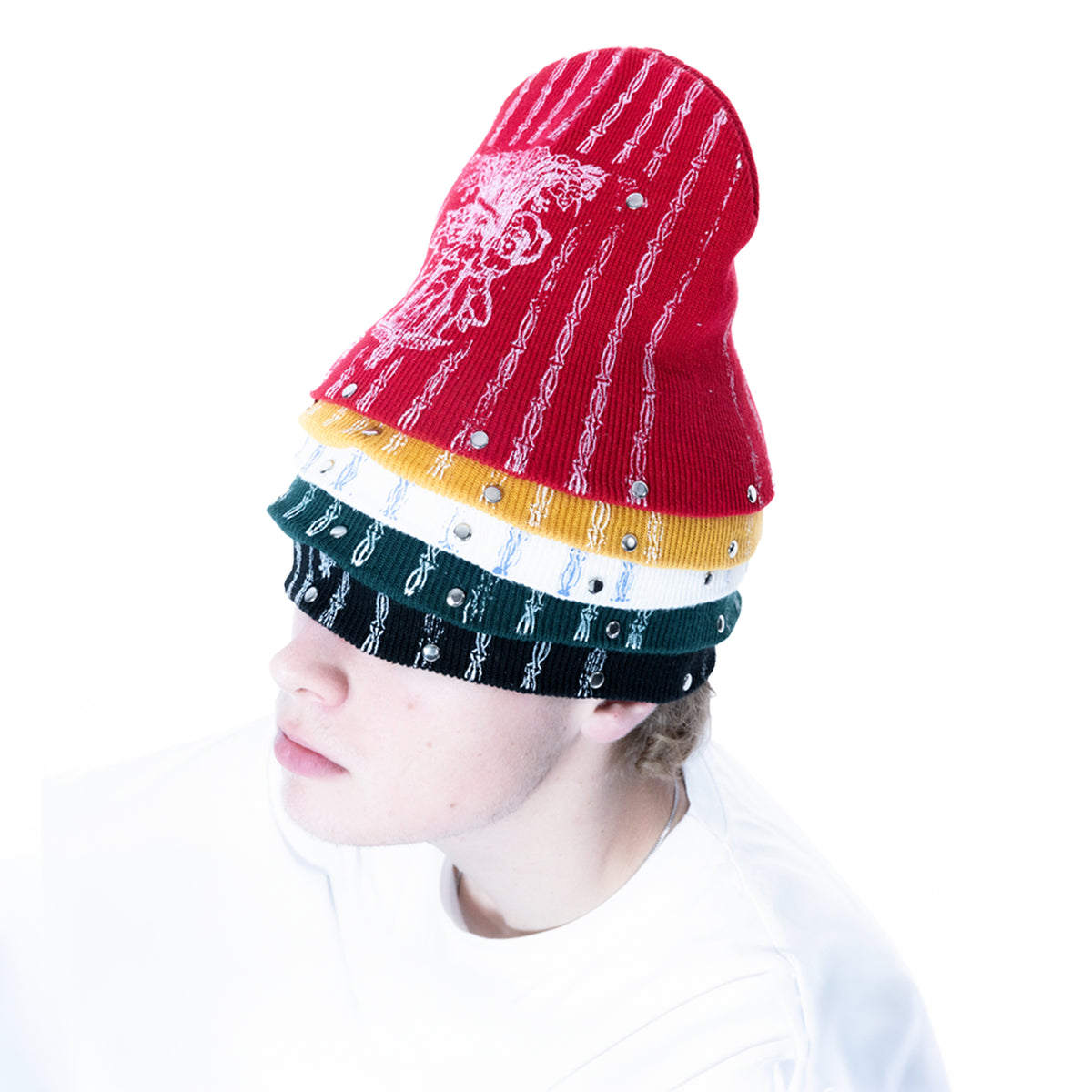 A person shown in profile wearing a tall knit beanie with horizontal color bands: red on top, then mustard yellow, white, dark green, and black. The beanie is embellished with small silver rhinestones and has a white graphic design on the red section.
